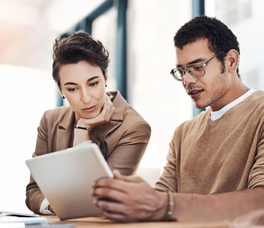 A woman and a man sitting at a table, looking at a tablet together in a bright room.