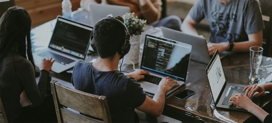 People working on laptops at a wooden table, some with headphones, in a casual setting.