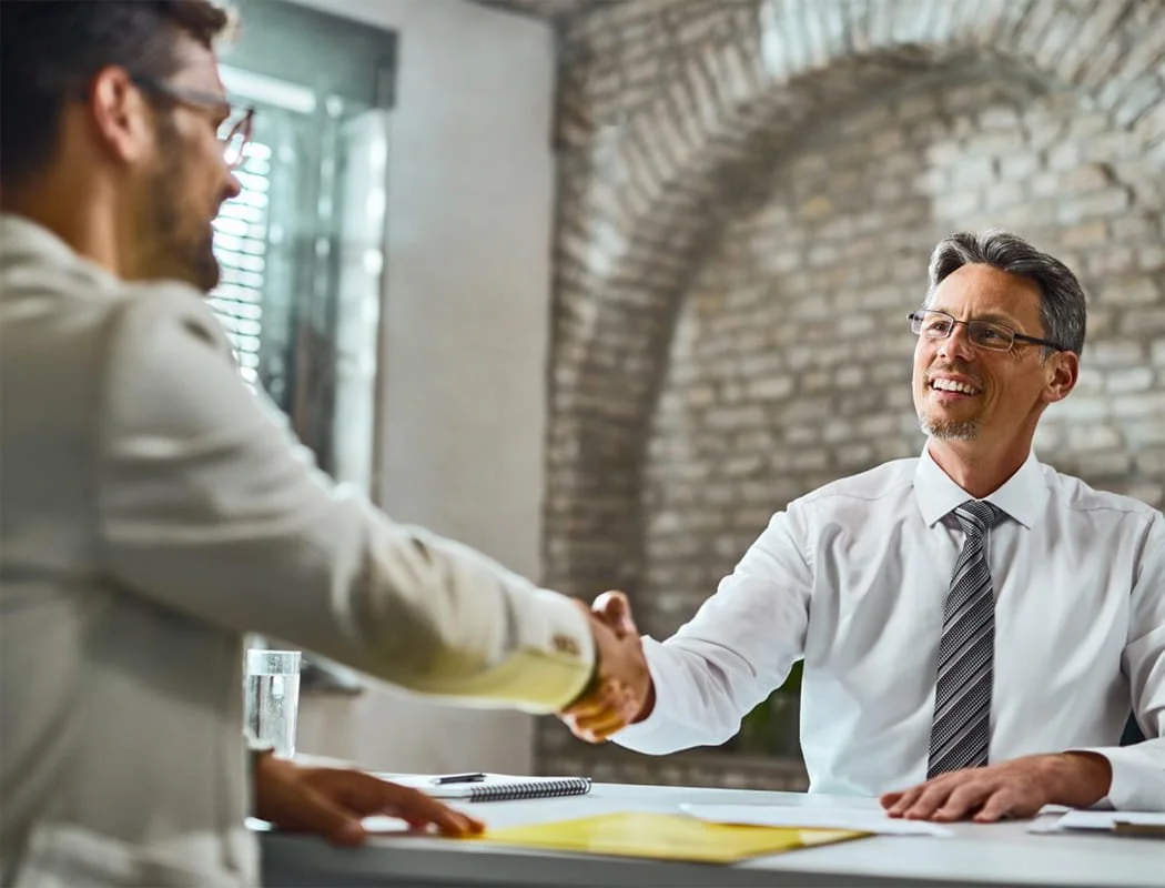 Two men shaking hands across a desk in a modern office with a brick wall and window in the background.
