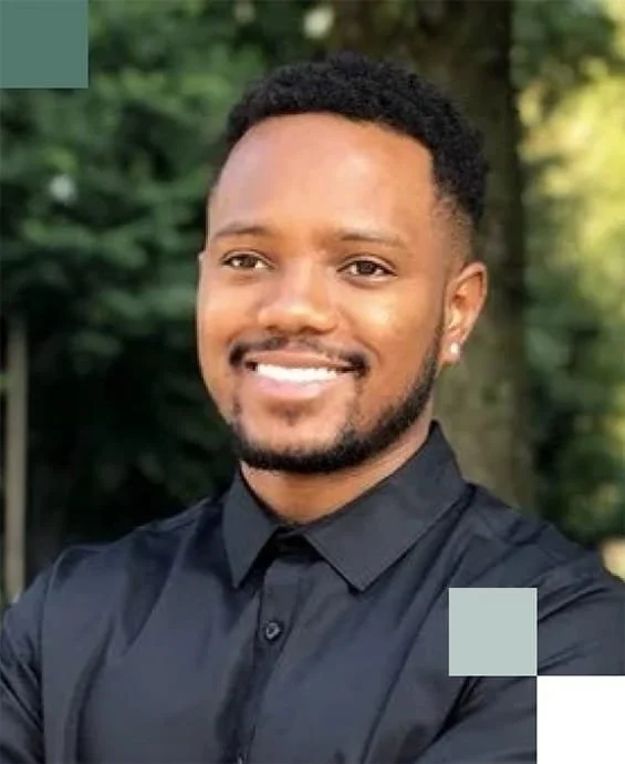 A smiling man with short curly hair, a beard, and earrings, wearing a dark button-up shirt, standing outdoors with trees in the background.
