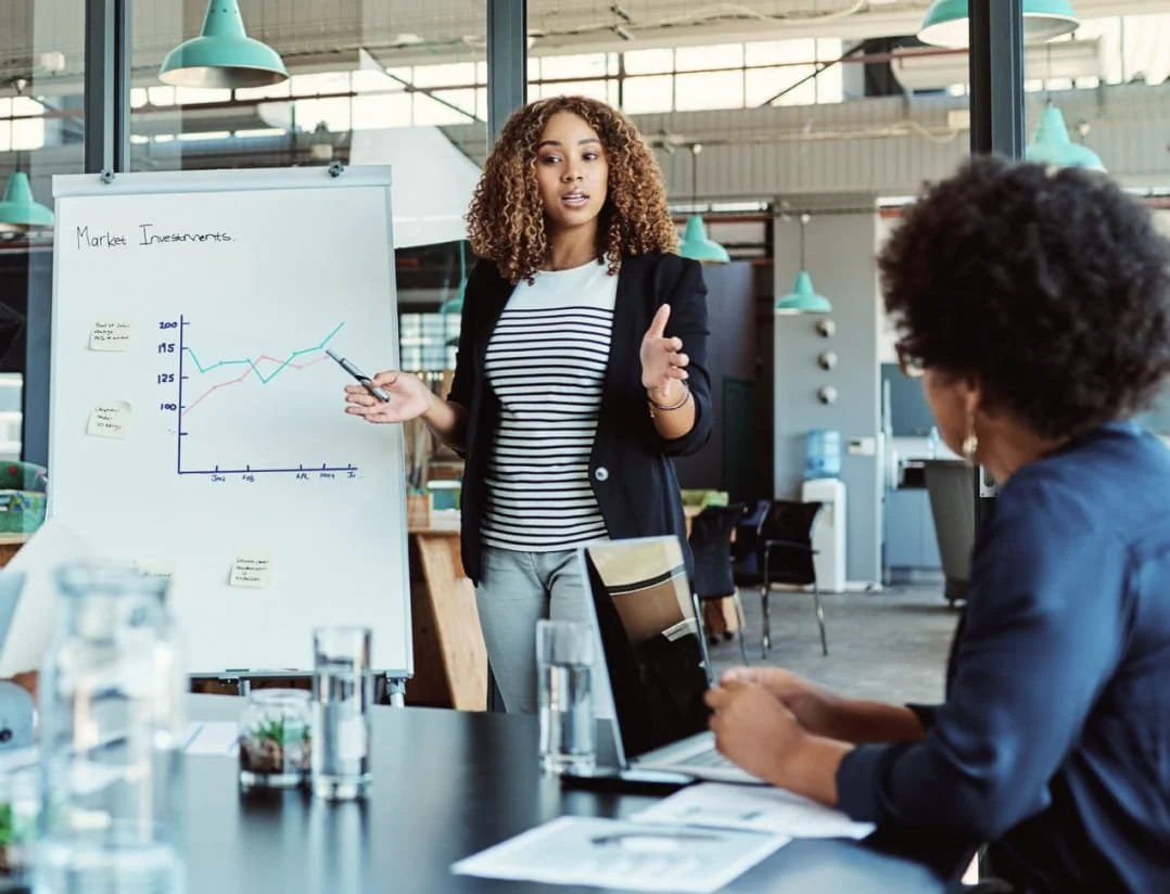 A woman presenting market investments data on a whiteboard to colleagues in a modern office conference room.