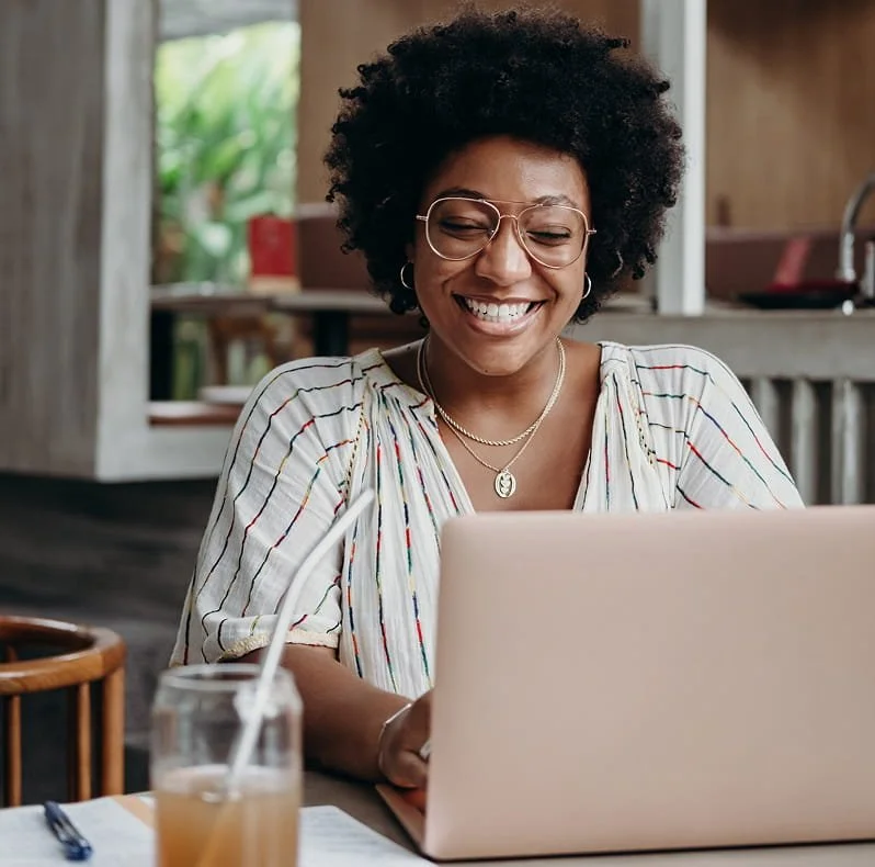 A woman with curly hair and glasses smiling while working on a laptop at a table with a notebook, pen, and glass of iced tea.