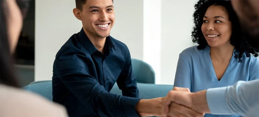A man in a blue shirt shaking hands with another person in an office setting, while two women look on and smile.