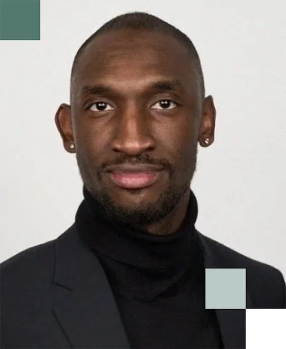 Portrait of a man with dark skin, wearing a black turtleneck and a dark blazer, with short hair and earrings, against a plain white background.