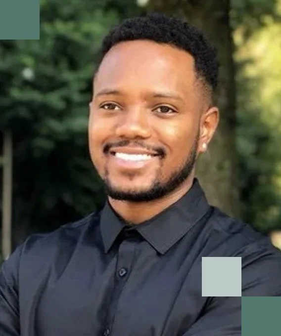 Young man with short curly hair and a beard smiling outdoors, wearing a black button-up shirt.