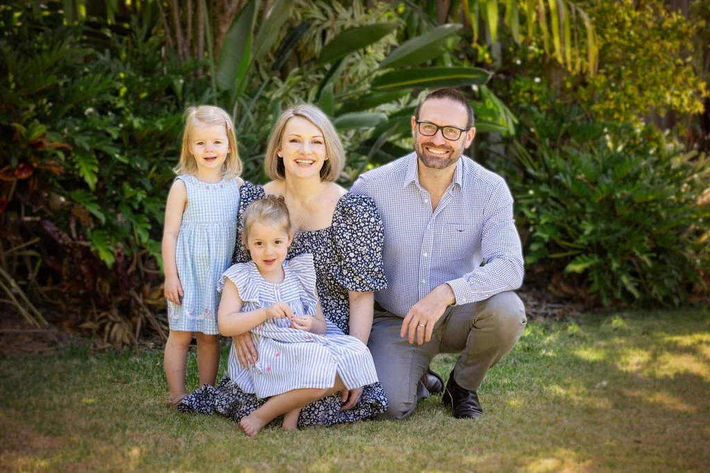 A happy family of four outdoors, with a woman, a man, and two young girls, posing in front of lush green plants.