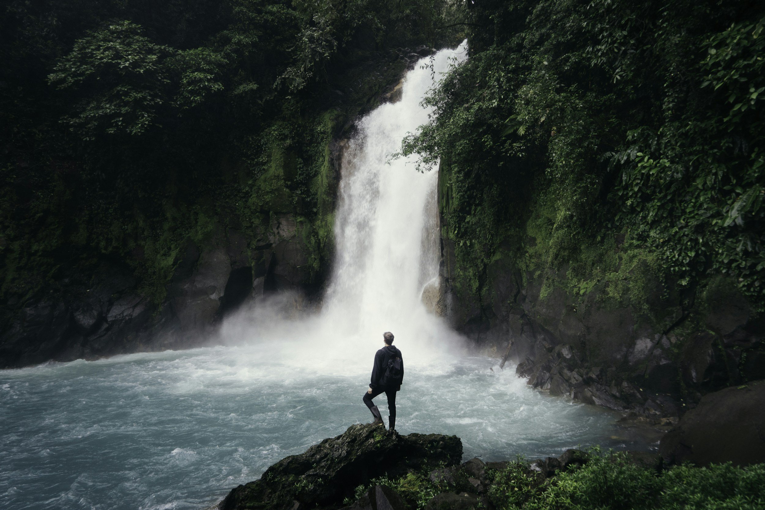 A person standing on a large rock, facing a waterfall surrounded by lush green foliage.