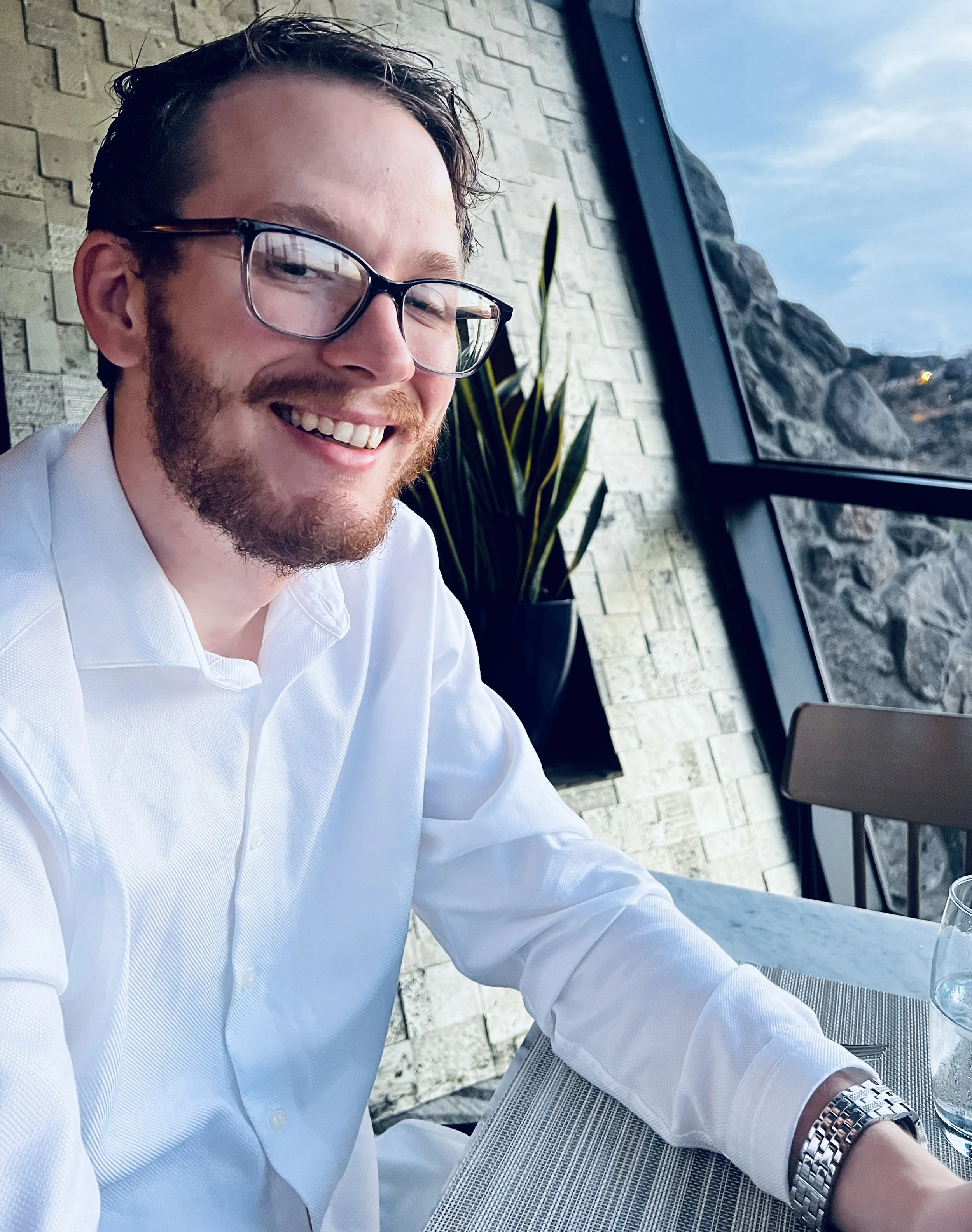 A man with glasses and a beard smiling while sitting at a table in a restaurant, with a stone wall, potted plant, and large window showing rocks and sky in the background.