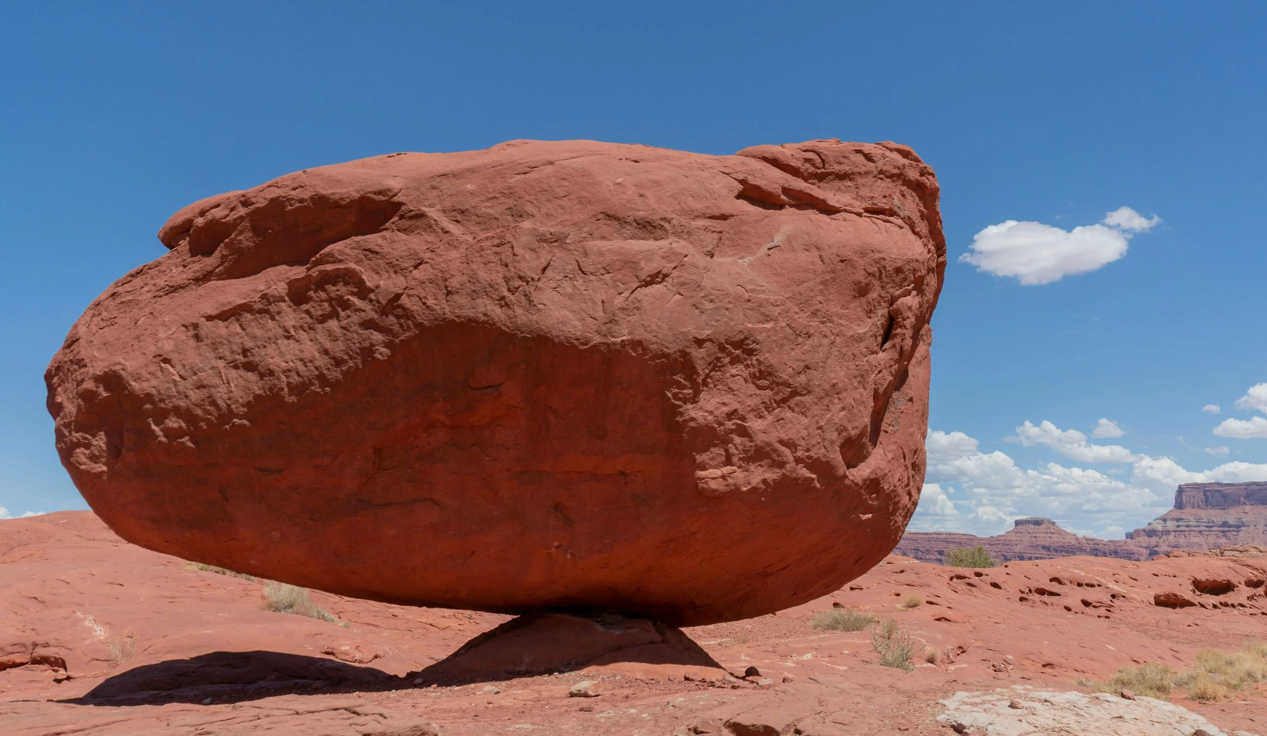 A large red sandstone boulder balanced on a small rock in a desert landscape with a blue sky and a few scattered clouds.