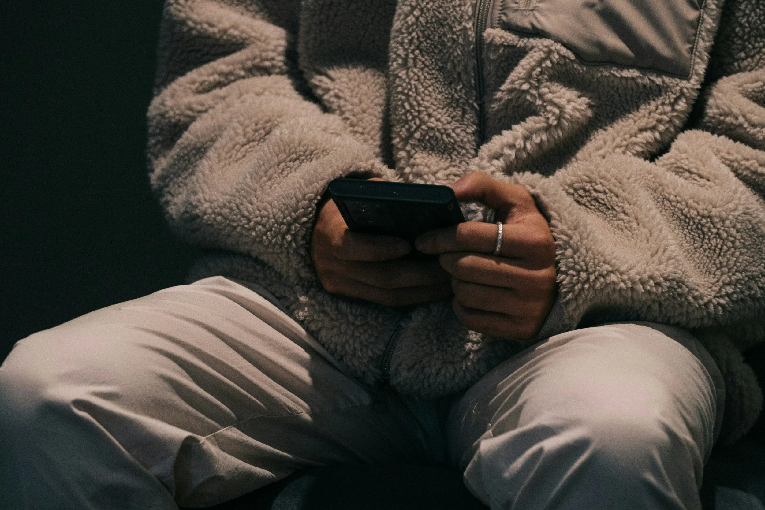Person sitting on a dark surface wearing a beige fleece jacket and light-colored pants, holding and using a smartphone.