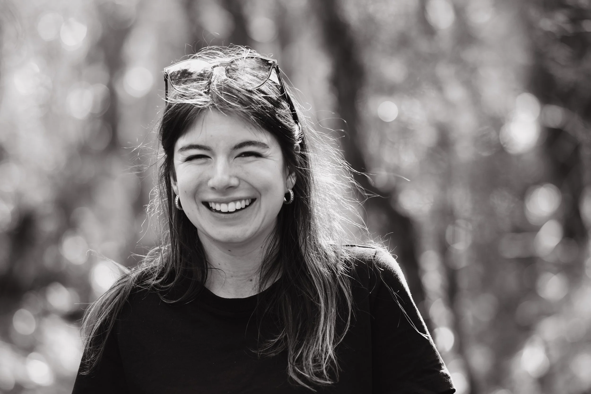 Black and white photo of a woman smiling outdoors, with sunglasses on her head and blurred trees in the background.