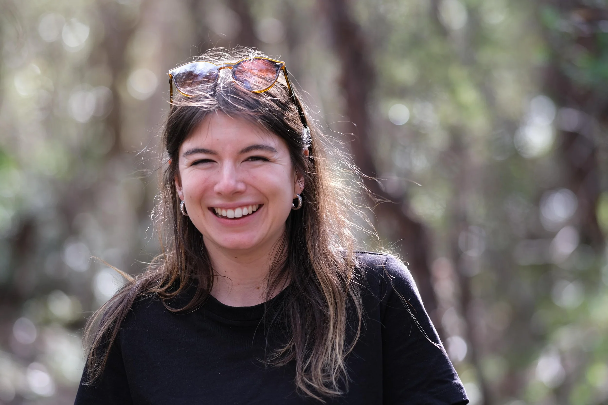 A smiling young woman outdoors in a wooded area, wearing sunglasses on her head and a black top.