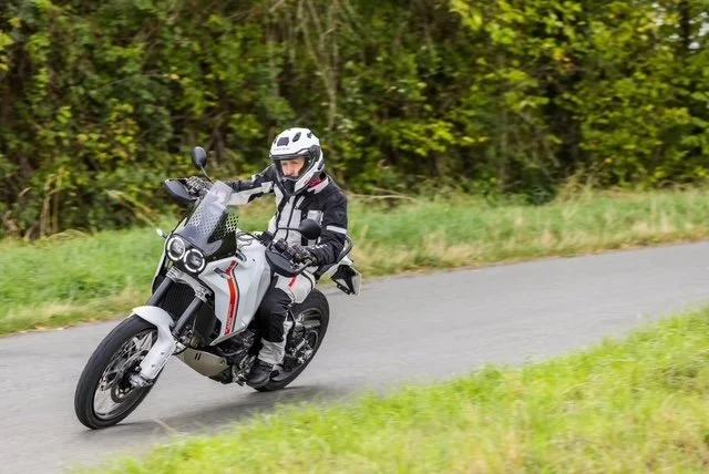 Motorcycle rider wearing a helmet and protective gear riding on a paved road with trees in the background.