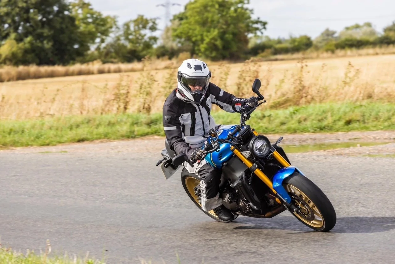 Motorcycle rider wearing a helmet and riding on a curved paved road with trees and field in the background.