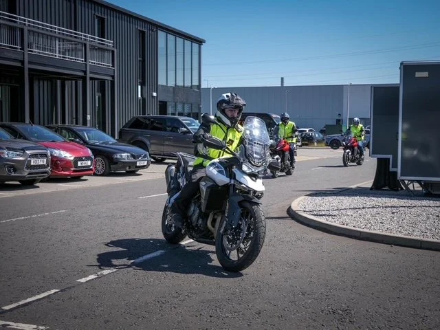 Police officers riding motorcycles in a parking lot with cars and a modern building in the background on a sunny day.