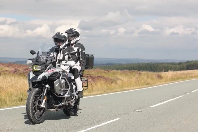 Two motorcyclists riding a gray adventure motorcycle on a rural road with open fields and cloudy skies in the background.