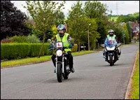 Two police officers riding motorcycles on a suburban street with green trees and houses in the background.