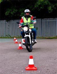 A person riding a motorcycle through an obstacle course marked by orange traffic cones, wearing a helmet and safety vest.