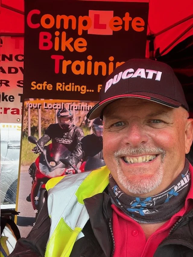 A smiling man wearing a Ducati cap, a high-visibility vest, and a neck gaiter pose in front of a sign that reads 'Complete Bike Training' with the tagline 'Safe Riding...'. There are motorcyclists pictured on the sign in the background.