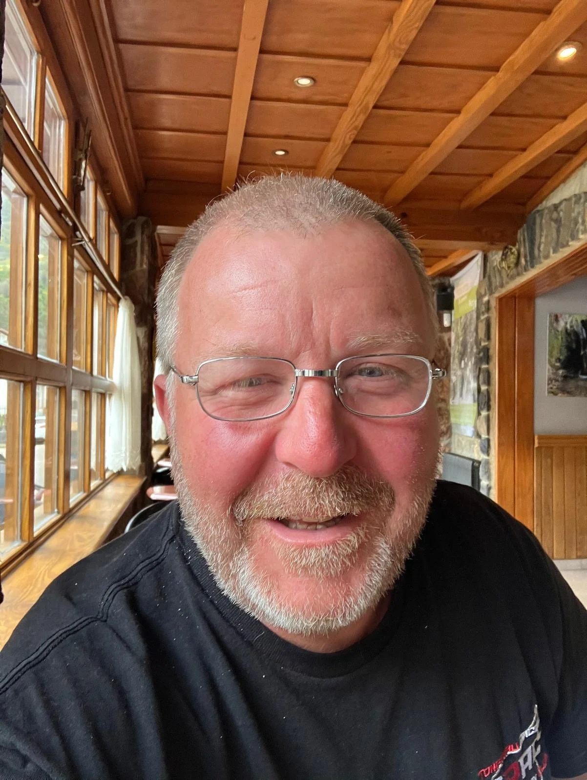 A smiling man wearing glasses with a beard and short hair, inside a rustic wooden cabin or restaurant with large windows, wooden ceiling beams, and stone walls.