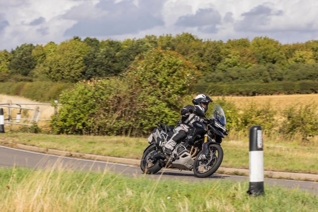 Motorcyclist riding a black motorcycle along a rural road with greenery and fields in the background.