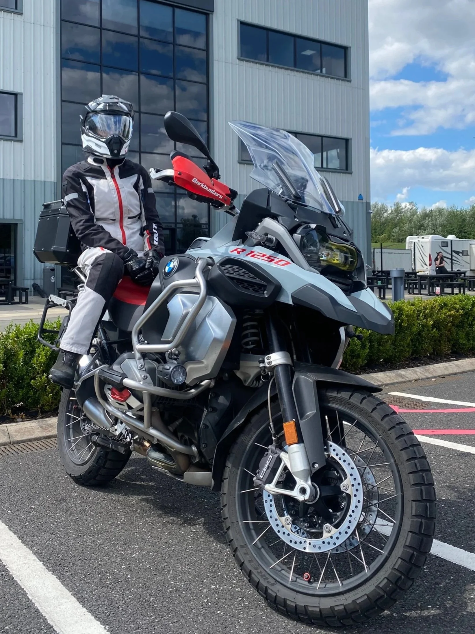 A person in motorcycle gear sitting on a BMW R 1250 GS adventure motorcycle, parked outside a building with large windows and a blue sky with clouds in the background.