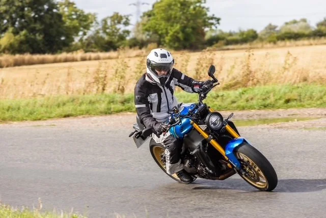 A motorcyclist wearing a helmet and protective gear riding a blue and black motorcycle on a curved road with grassy fields and trees in the background.