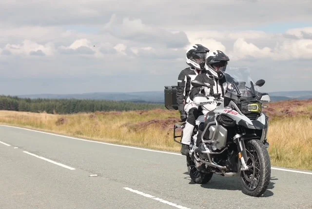 Two motorcyclists in gear riding a motorcycle on a rural road with open fields and cloudy sky in the background.
