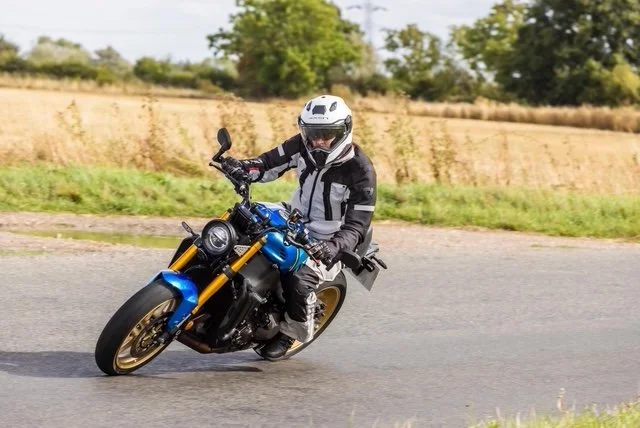 Motorcycle rider wearing a white helmet and riding gear leaning into a turn on a blue motorcycle on a paved road with grassy and wooded landscape in the background.