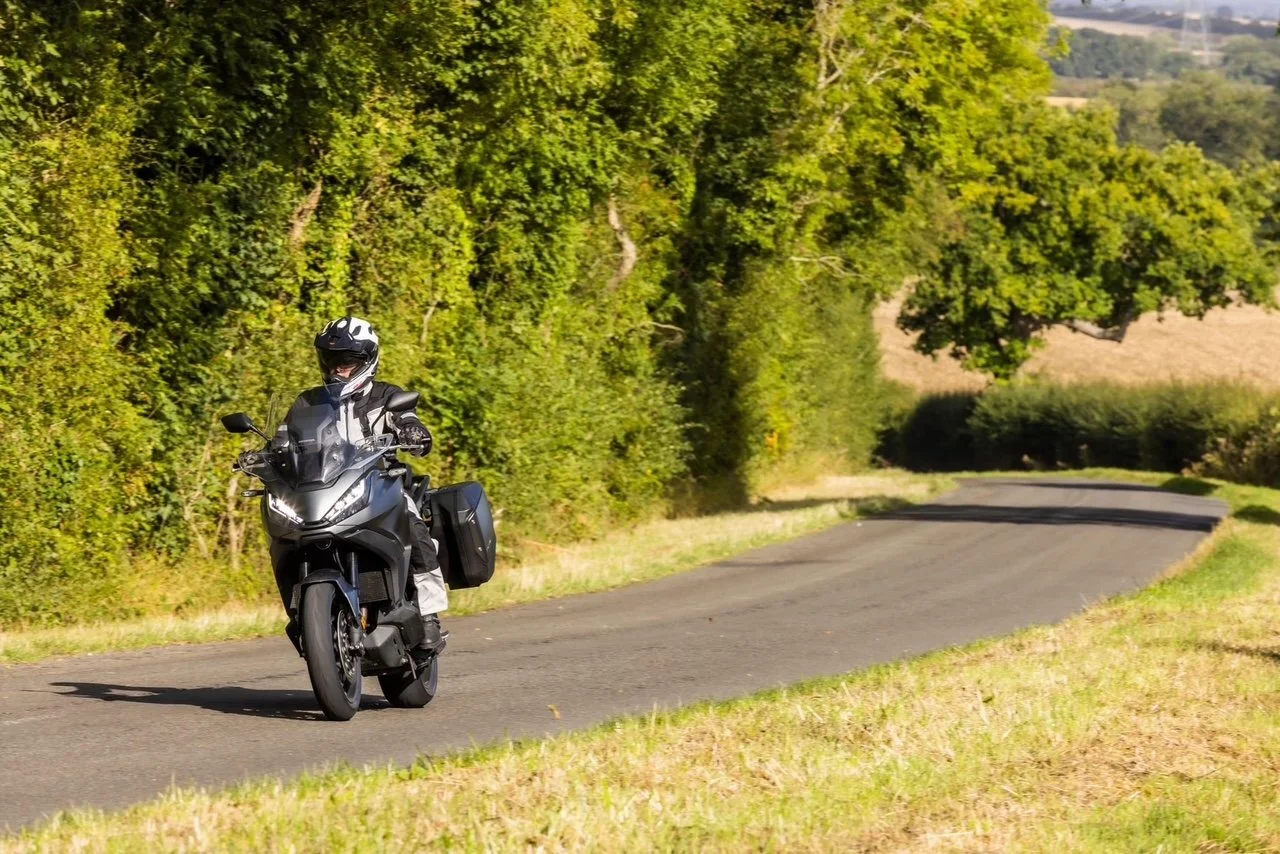 A person riding a black motorcycle along a curved rural road with lush green trees and fields in the background.