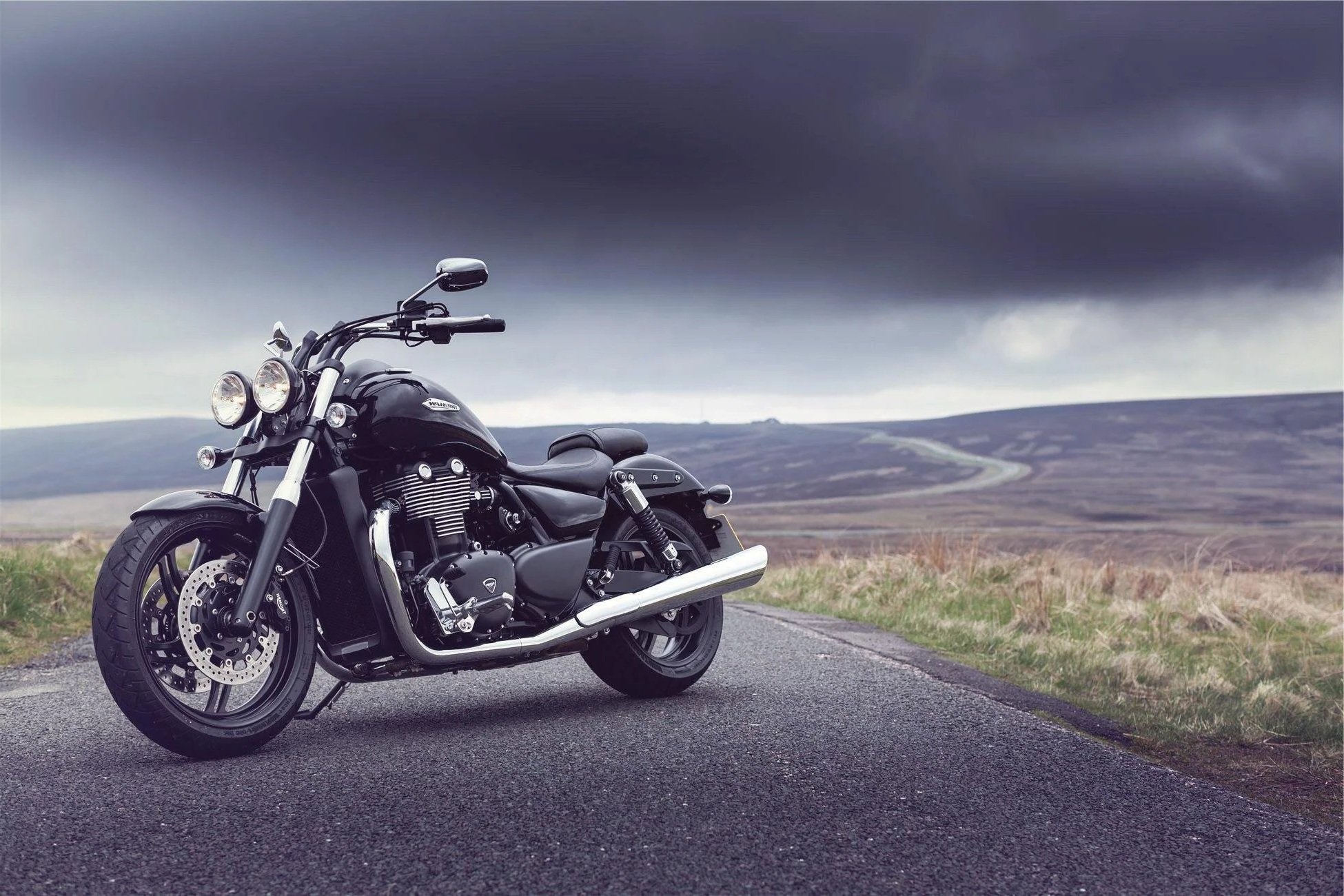 A black motorcycle parked on a rural road with rolling hills and dark stormy clouds in the background.