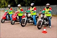 Four people in reflective safety vests standing next to motorcycles and scooters during a training session on a paved area with orange cones.