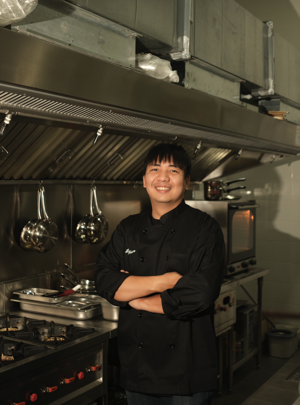 A smiling chef in a black uniform standing with arms crossed in a commercial kitchen with stainless steel cooking equipment and hanging ladles.