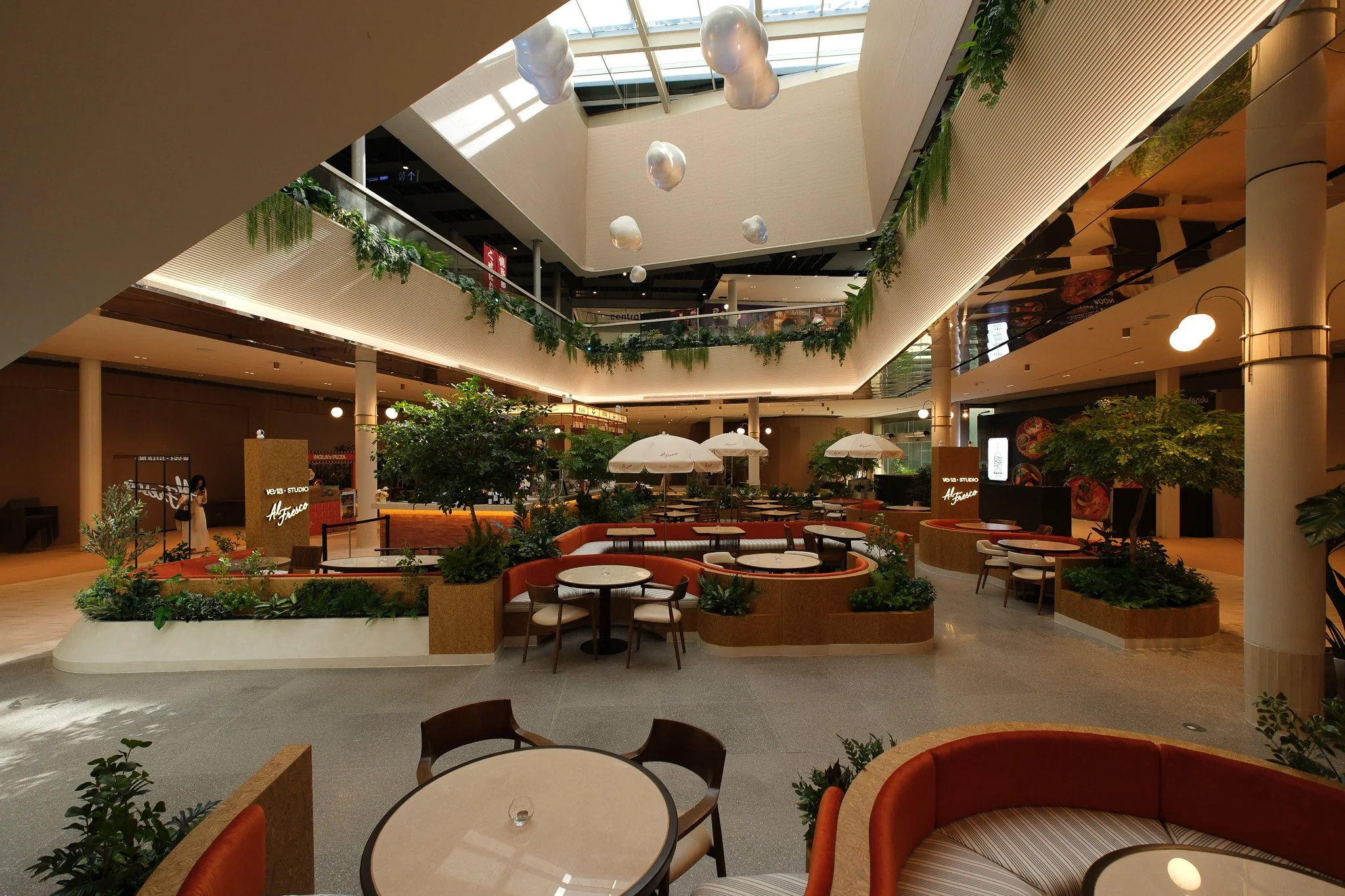 Interior view of a modern shopping mall or restaurant area with circular seating, green plants, hanging cloud-shaped light fixtures, and a glass ceiling allowing natural light to enter