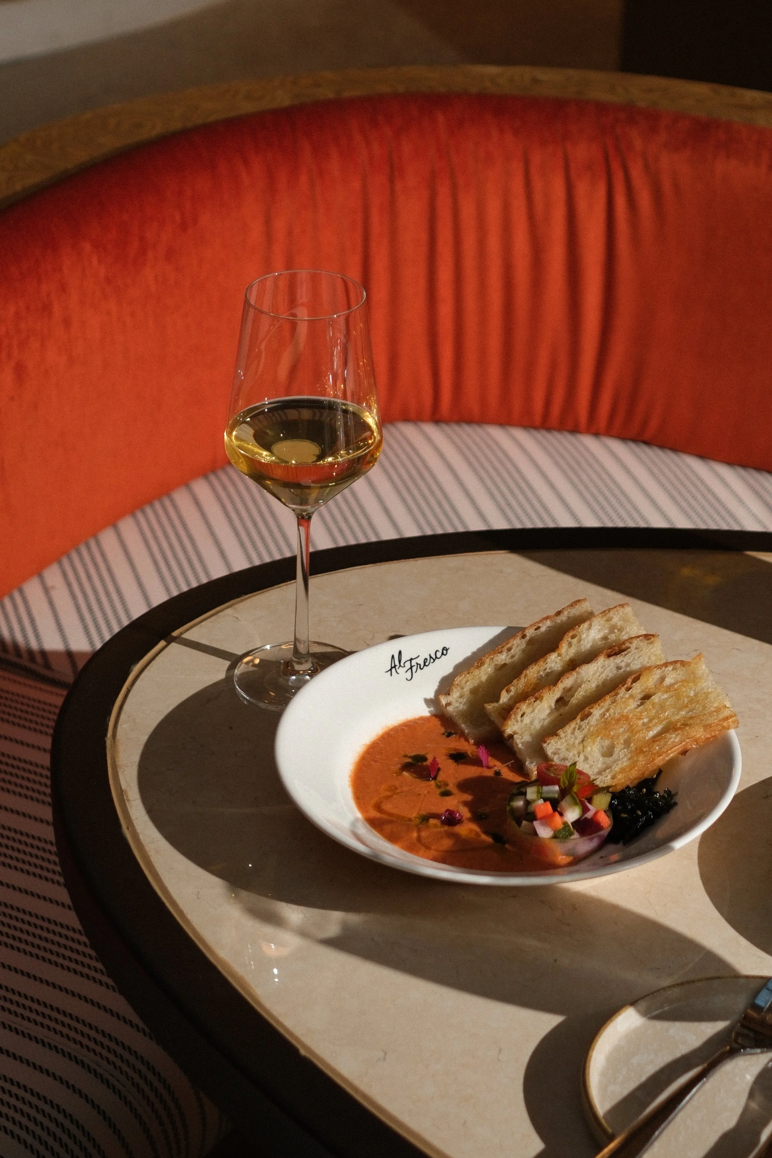 A glass of white wine and a bowl of tomato soup with bread slices on top, garnished with herbs and vegetables, on a small round table in a restaurant setting.
