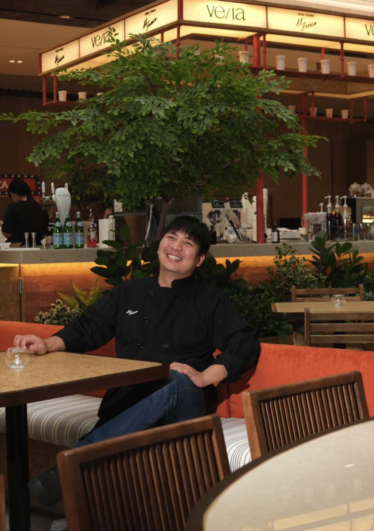 A smiling man wearing a black chef's coat sitting at a table in a restaurant with a large green tree behind him.