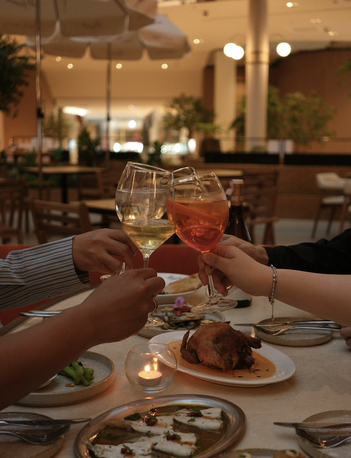 People celebrating with wine glasses at a dinner table in a restaurant, with dishes of food including chicken, seafood, and other appetizers, and a lit candle.
