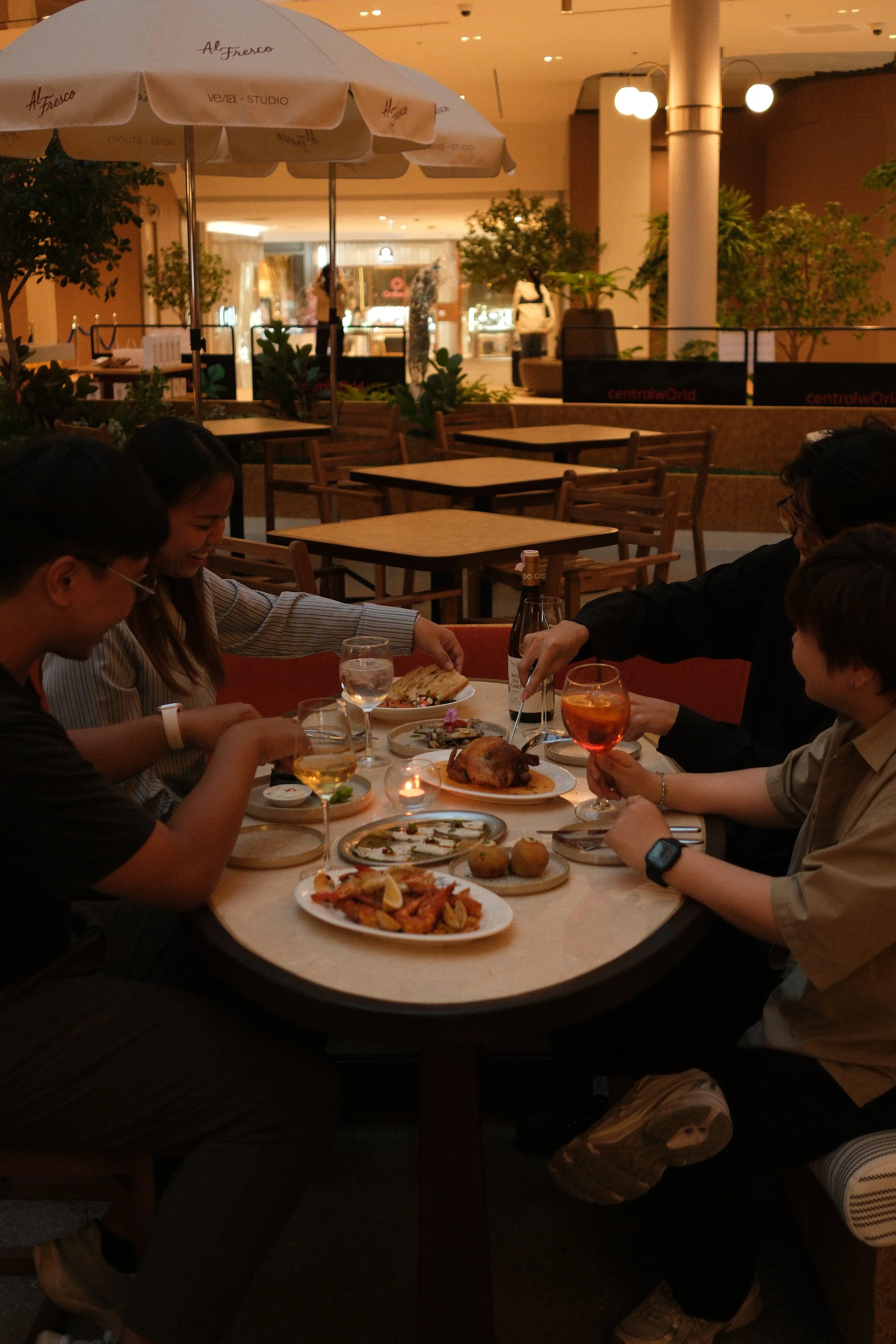 People enjoying a dinner at a restaurant table with various dishes, drinks, and a lit candle inside a shopping mall.