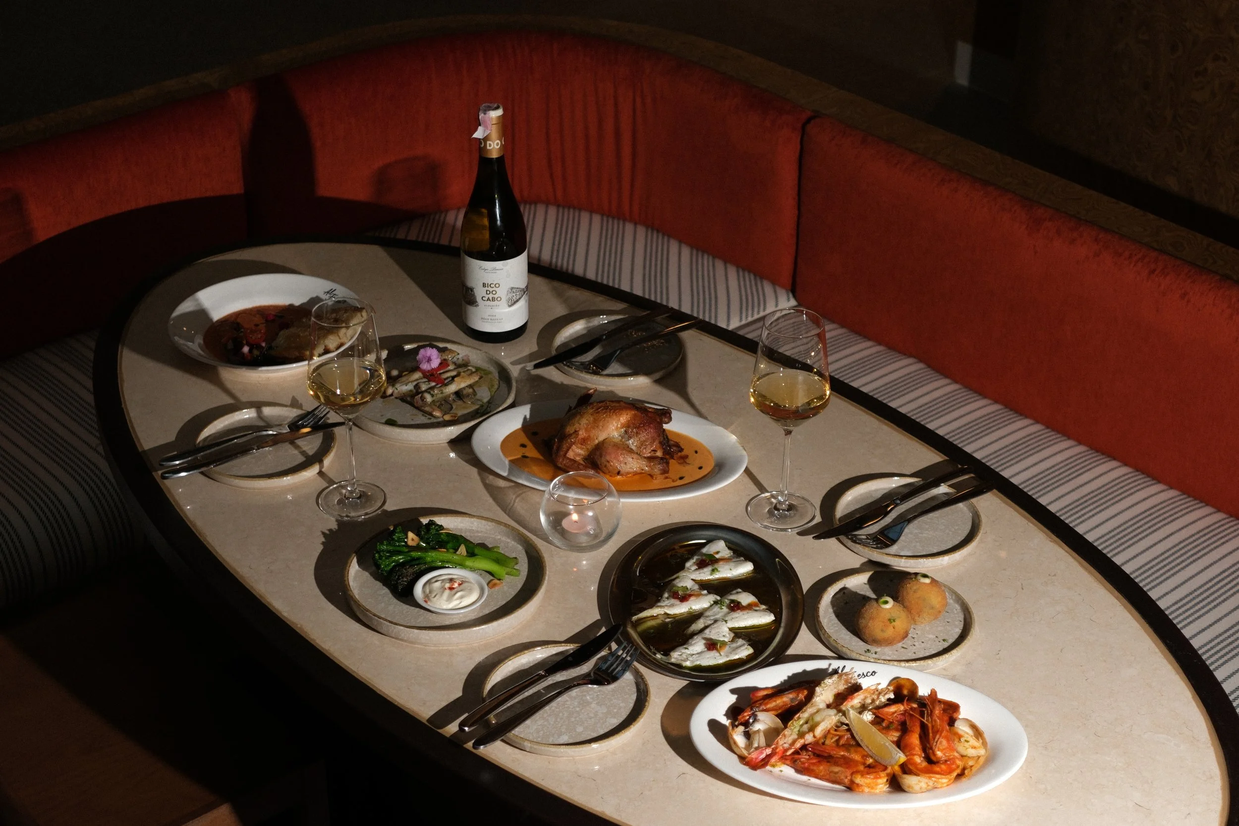Table with various plates of food, glasses of white wine, a bottle of wine, a candle, and utensils, in a dimly lit restaurant setting.