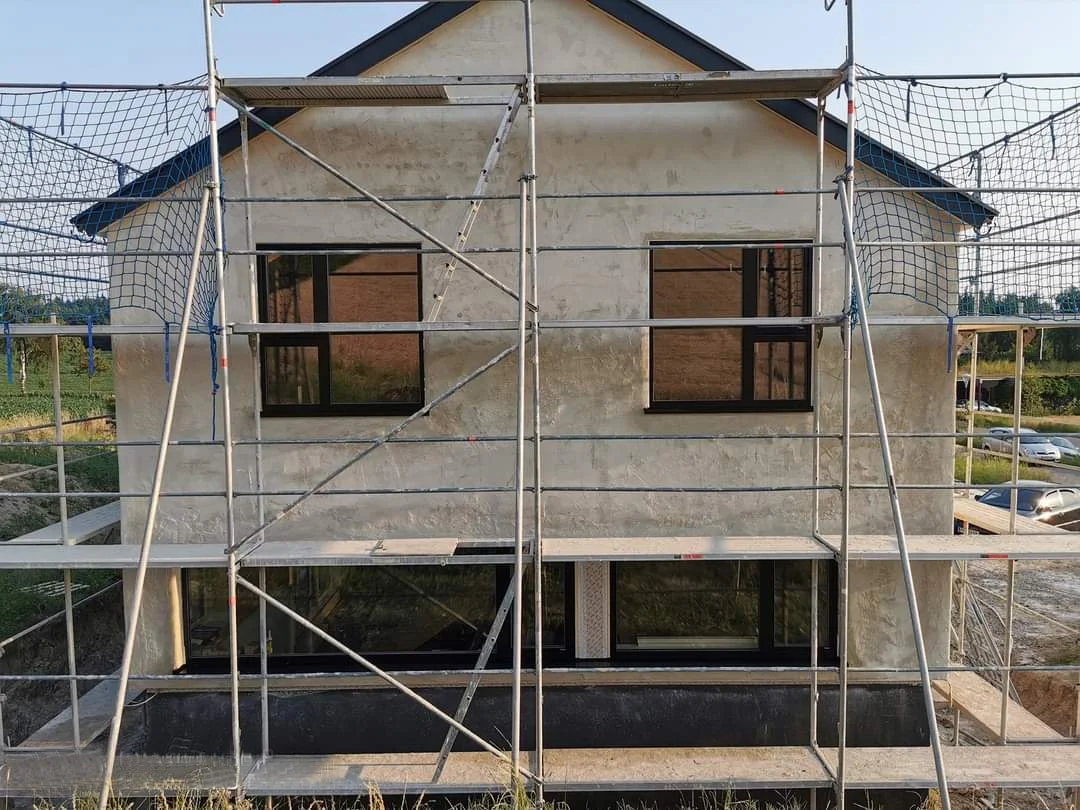 Ein Haus im Bau mit Gerüst, Fenster und Kalkfassade, im Hintergrund ein grüner Hügel.