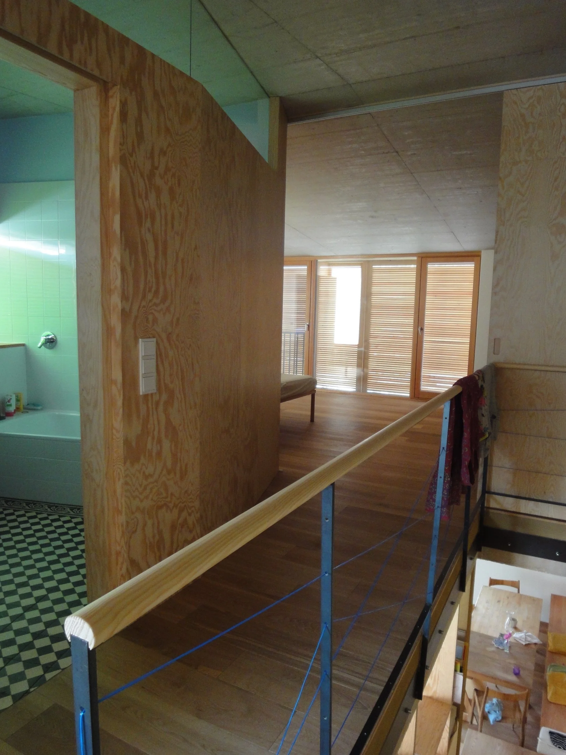 Blick auf eine offene Holzlamellentür im Innenbereich eines Hauses, mit Blick auf einen Wohnraum mit Holzboden und große Fenster mit Holzjalousien, sichtbar vom Obergeschoss aus.