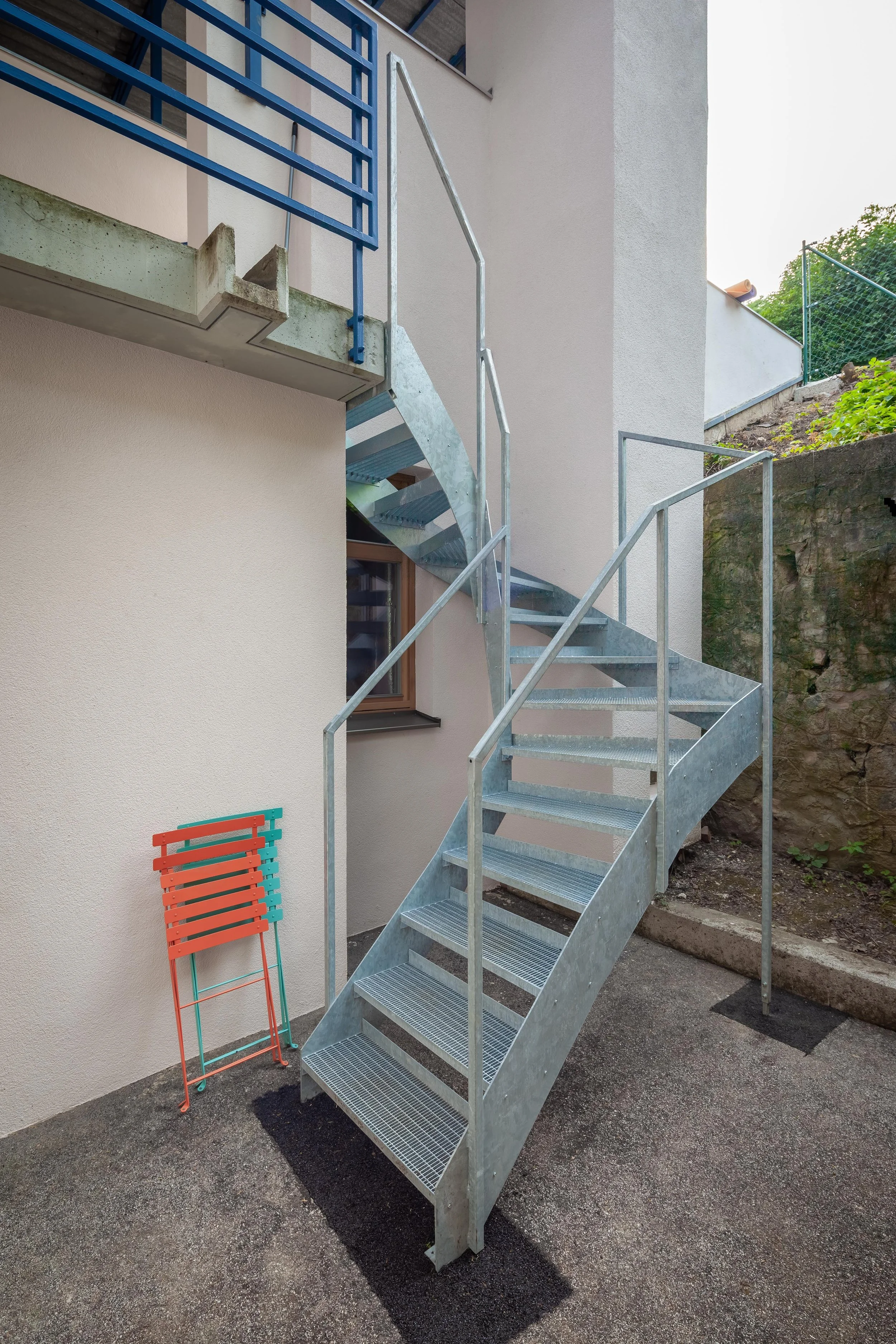Freitreppe aus Stahl vor einem Haus, daneben ein klappbarer Stuhl in Rot und Hellblau, im Hintergrund eine Mauer und ein Garten mit Bäumen.