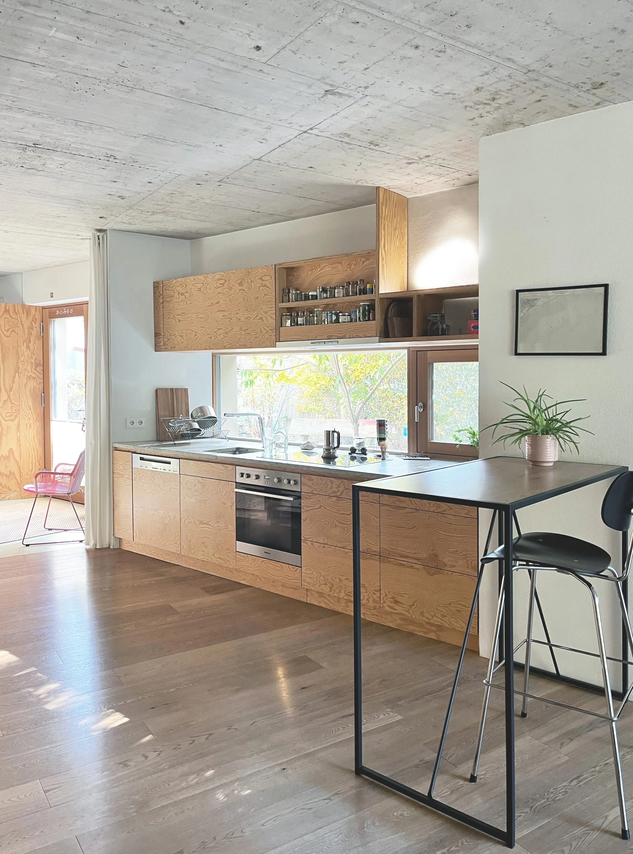 Moderne Küche mit Holzfronten, großen Fenster, Ofen, Herd, Spüle und Küchenutensilien, in einem hellen Raum mit Holzboden.