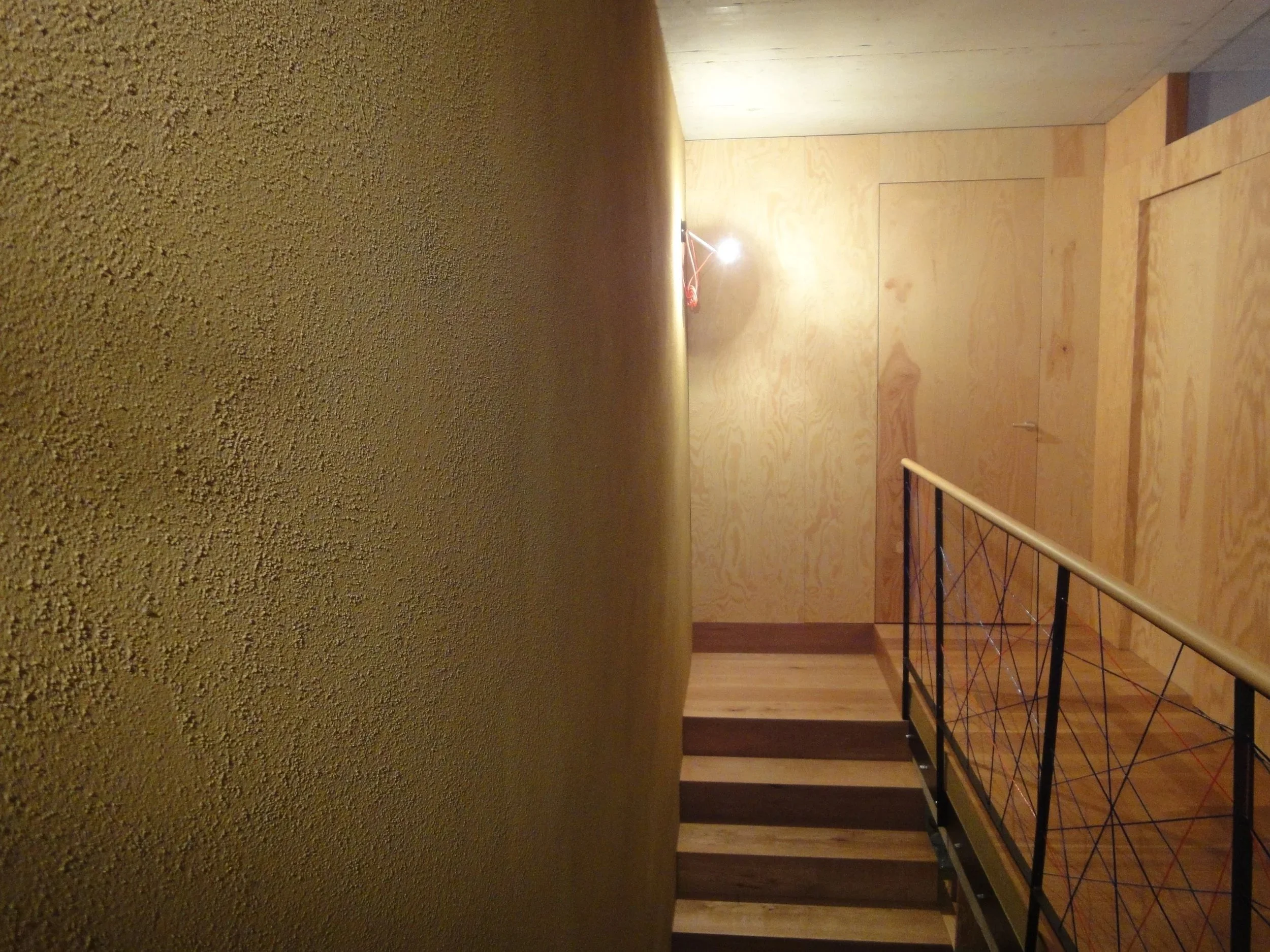 Innenaufnahme eines Treppenhauses mit Holzverkleidung, einer Handlaufstütze aus Metall, und einer einzelnen Glühbirne an der Wand, die leuchtet.