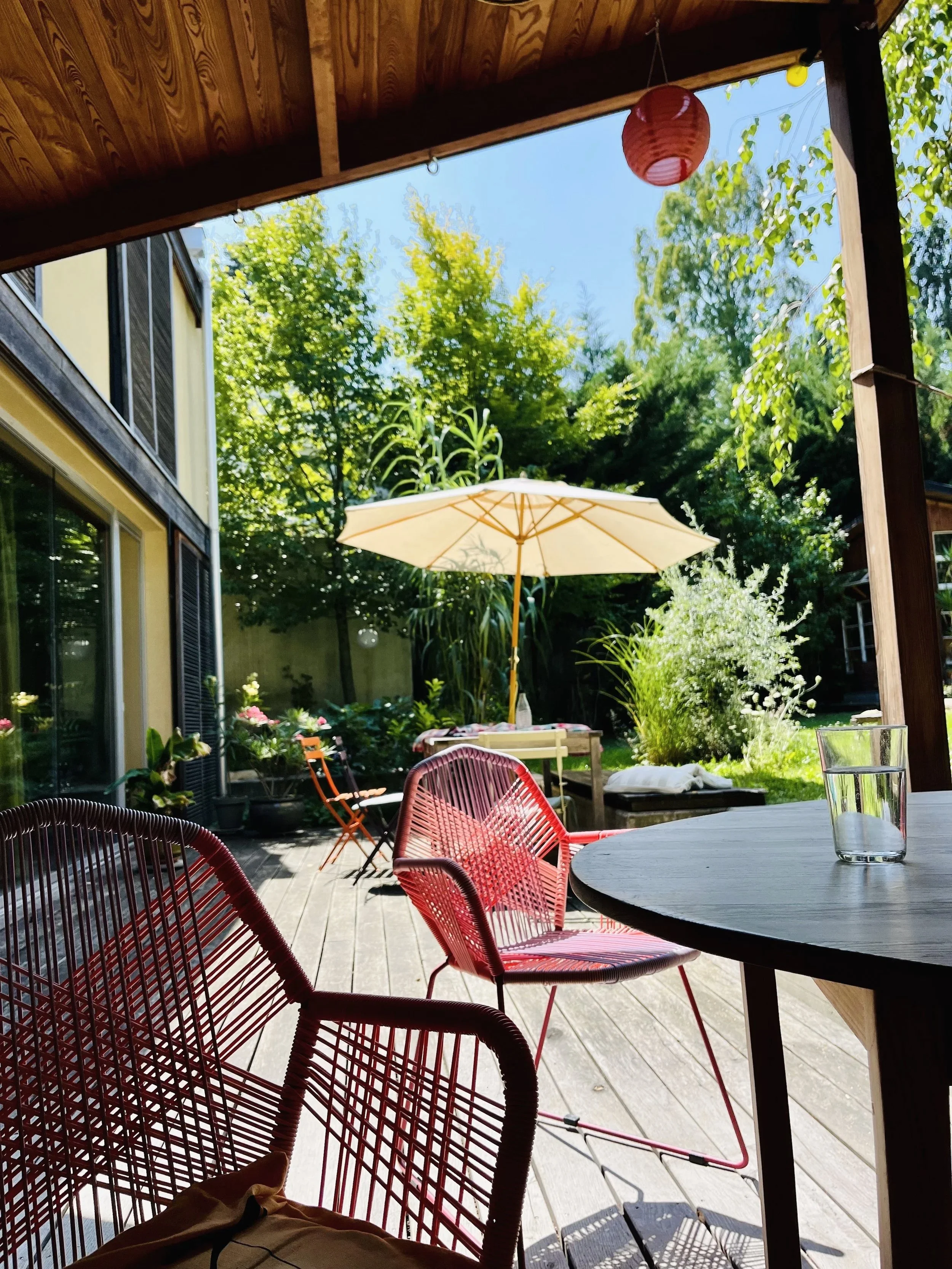 Blick aus einer überdachten Terrasse auf einen sonnigen Garten mit einem weißen Sonnenschirm, grünen Bäumen und bequemen Gartenmöbeln.