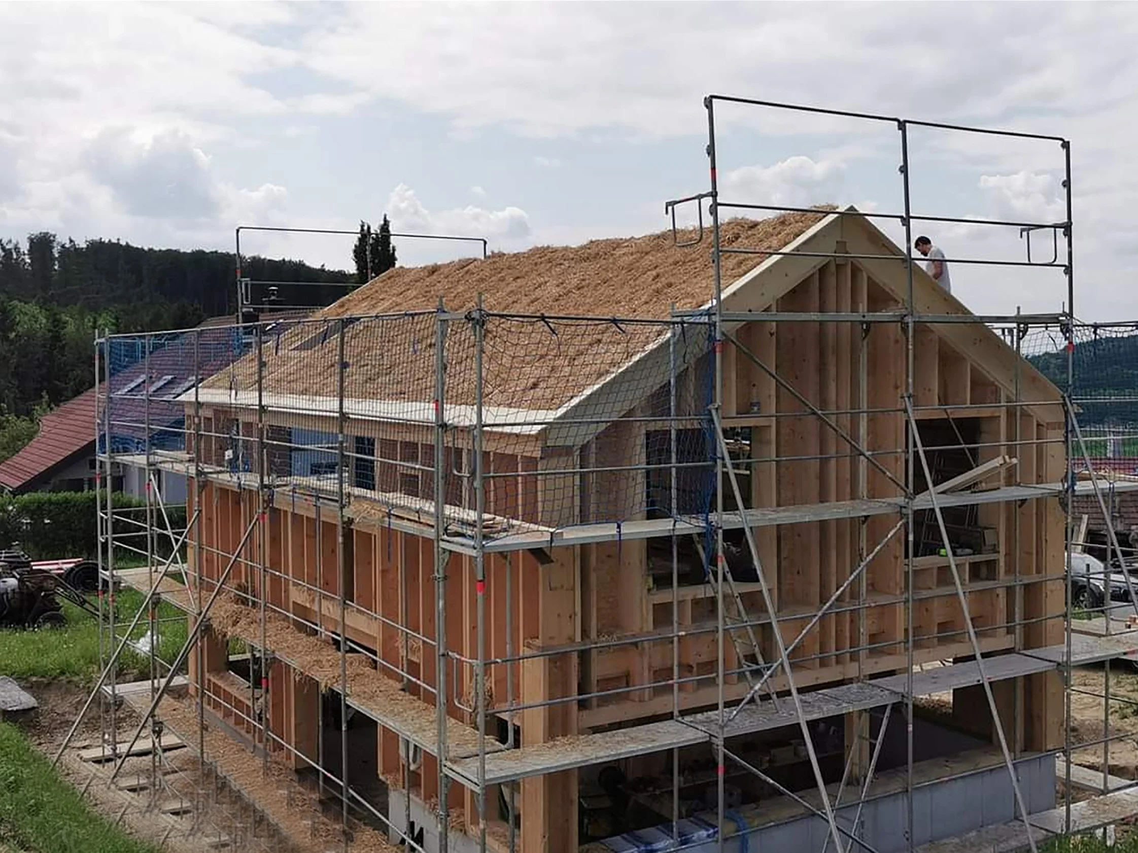Ein im Bau befindliches Holzhaus mit Strohdach, auf dem Dach wird gearbeitet, im Hintergrund Wald und ein bewölkter Himmel.