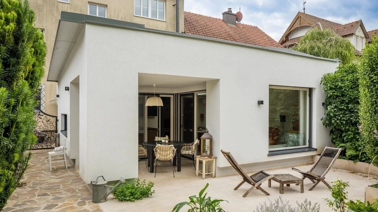 Ein modernes, weißes Haus mit großer Fensterfront und kleiner Veranda, im Außenbereich liegen zwei Liegestühle und ein Tisch, umgeben von Grünpflanzen und Bäumen.