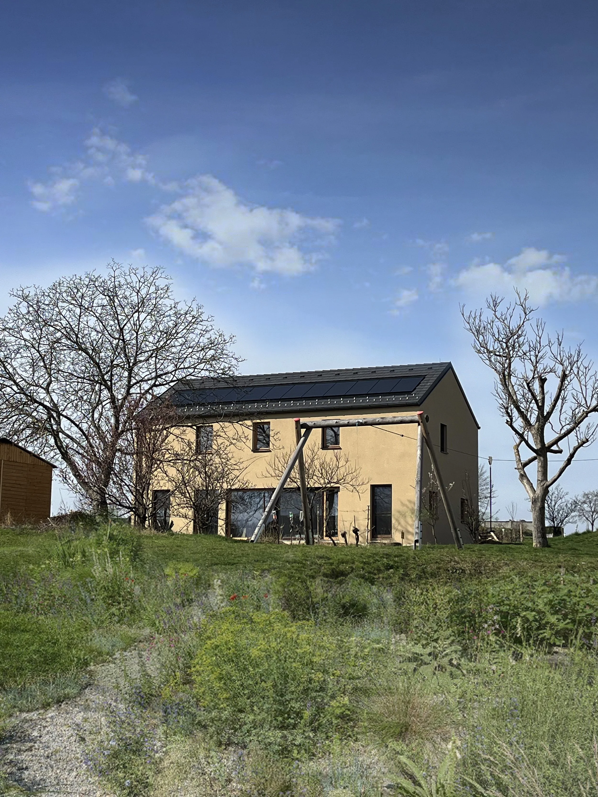 Neues Haus mit Solaranlagen auf dem Dach, umgeben von Bäumen und grünem Garten, bei sonnigem Himmel.