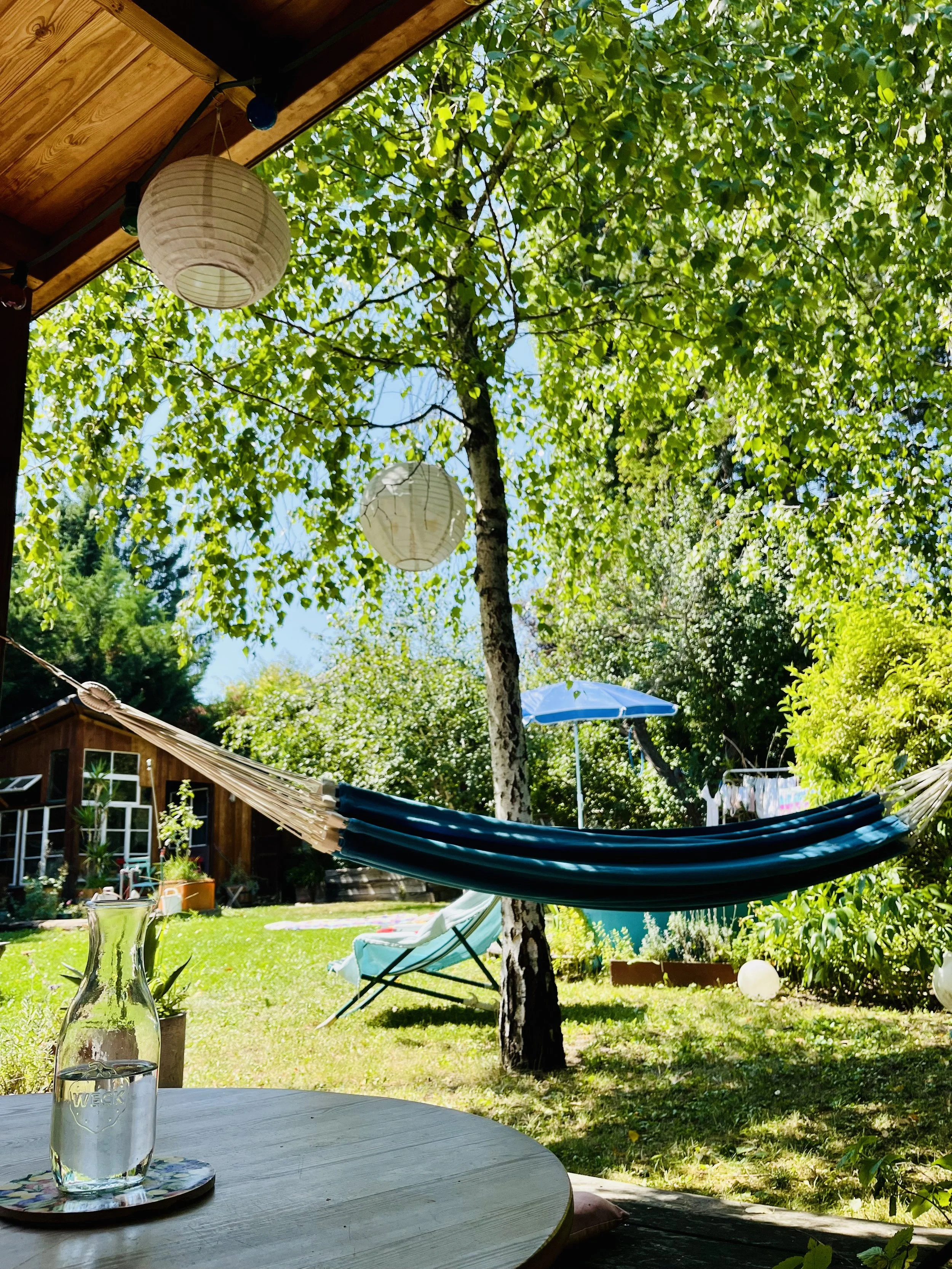 Garten mit Hängematte, Sonnenschirm, Liegestuhl, Bäumen und einem kleinen Gartenhaus, sonniger Tag.