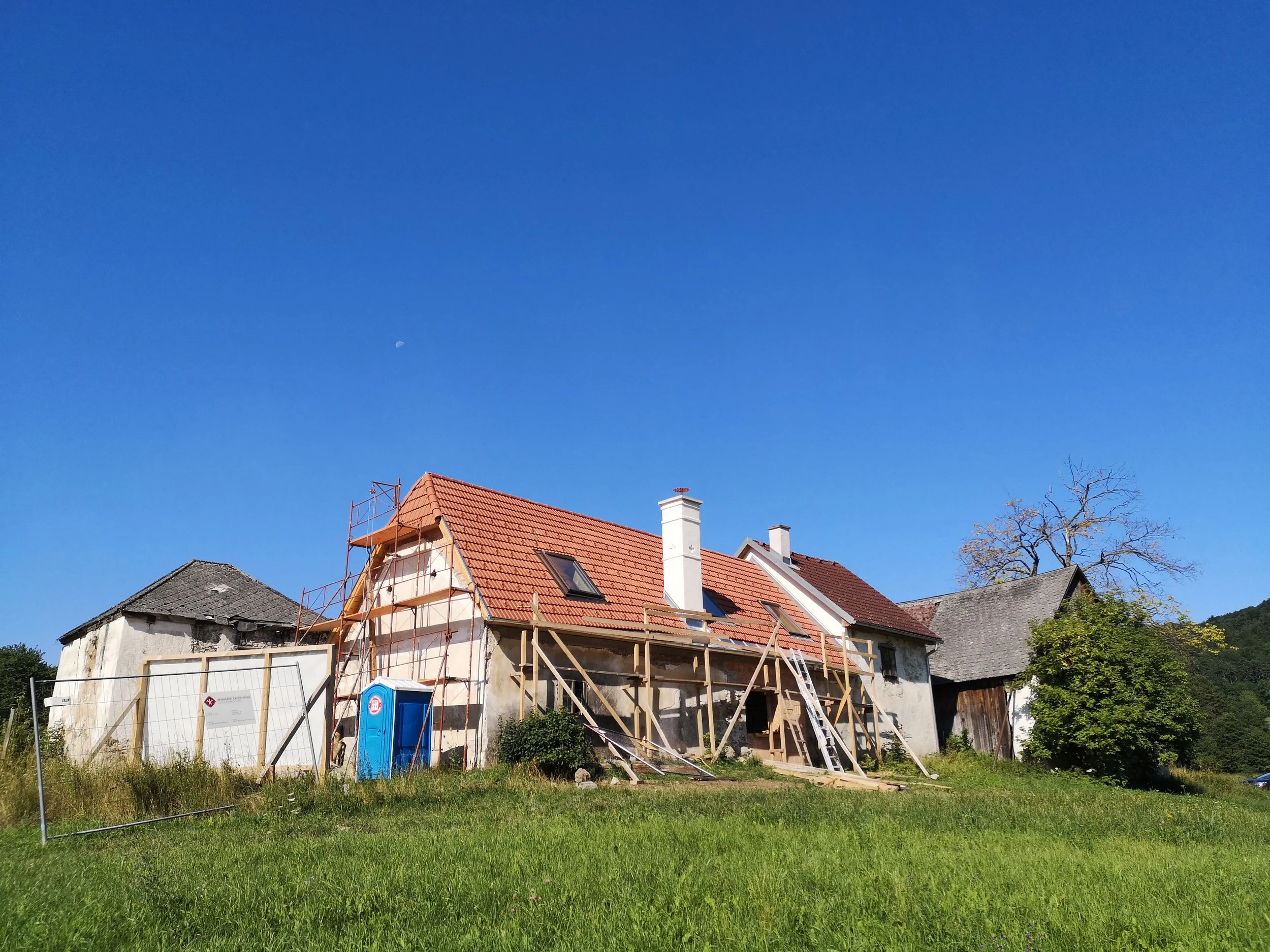 Ein Haus wird renoviert, mit Bauzäunen und einem Baugerüst, im Hintergrund ein blauer Himmel und Bäume.
